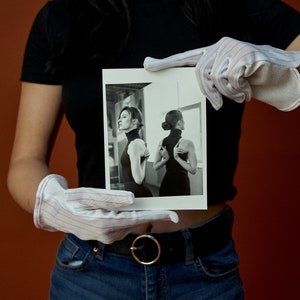May include: A black and white photograph held by gloved hands. The photograph features a woman in a black turtleneck dress, with one image showing her profile and another her back. The background is a neutral color.