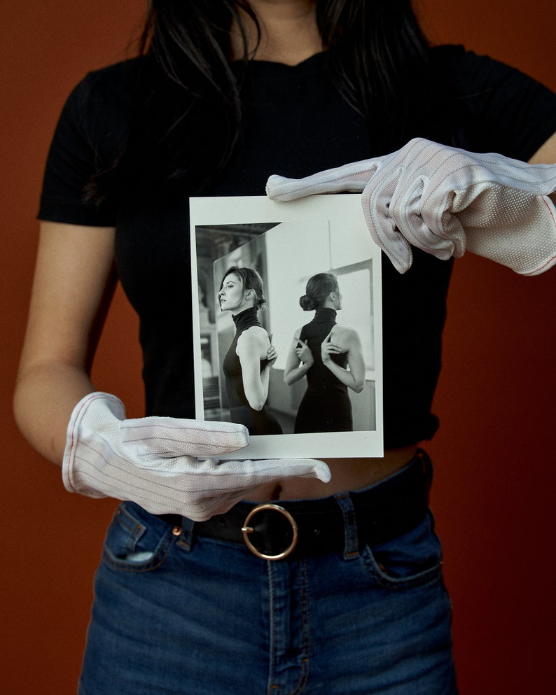 May include: A black and white photograph held by gloved hands. The photograph features a woman in a black turtleneck dress, with one image showing her profile and another her back. The background is a neutral color.