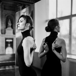 May include: A black and white photograph of a person in a black sleeveless turtleneck dress, standing in front of a mirror. The person's arms are crossed behind their back. A reflection of the person is visible in the mirror.
