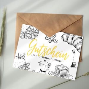 Könnte beinhalten: Ein weißer Gutschein mit einem braunen Umschlag. Der Gutschein zeigt Schwarz-Weiß-Illustrationen von Frühstücksartikeln wie Kaffeebohnen, einem Croissant und Pfannkuchen. Der Text "Gutschein" ist gelb.