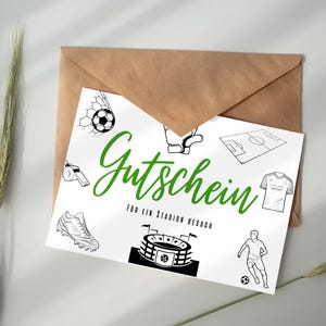 Puede incluir: Un vale de regalo blanco con texto verde e ilustraciones de fútbol, ​​incluido un balón de fútbol, ​​un estadio y un jugador, descansa sobre un sobre marrón. El texto dice "Gutschein" y "FÜR EIN STADION BESUCH".