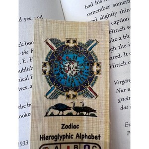 May include: A vintage-style bookmark featuring a blue and gold zodiac wheel surrounded by hieroglyphs. The bookmark is made of papyrus-like paper and features two black birds at the bottom. The text on the bookmark reads "Zodiac" and "Hieroglyphic Alphabet".