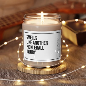 May include: A brown candle with a white label that reads "Smells Like Another Pickleball Injury". The candle is lit and sitting on a wooden coaster.