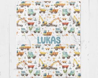 Op de afbeelding: Witte babydeken met een herhalend patroon van bouwmachines, sterren en teddyberen in geel, groen en oranje. De naam "LUKAS" is in blauw over het midden van de deken gedrukt.