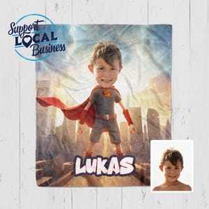 May include: A personalized superhero blanket featuring a child's face superimposed on a superhero body. The child is wearing a gray and red superhero outfit with a red cape. The name "LUKAS" is displayed at the bottom. The blanket has a cityscape background and the text "Support Local Business".