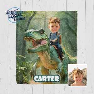 May include: A personalized blanket featuring a digitally created image of a child riding a green dinosaur in a forest setting. The child's name, "CARTER," is displayed in a green banner. The blanket also includes a small photo of the child and the text "Support Local Business."