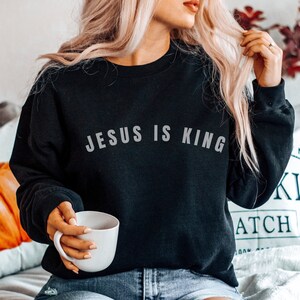 May include: A black sweatshirt with the text "JESUS IS KING" in white lettering. The sweatshirt is being worn by a person sitting on a couch.