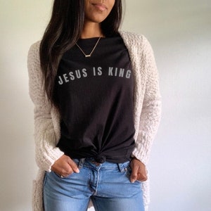 May include: A woman wearing a black t-shirt with the text "JESUS IS KING" in white lettering, a white cardigan, and blue jeans.