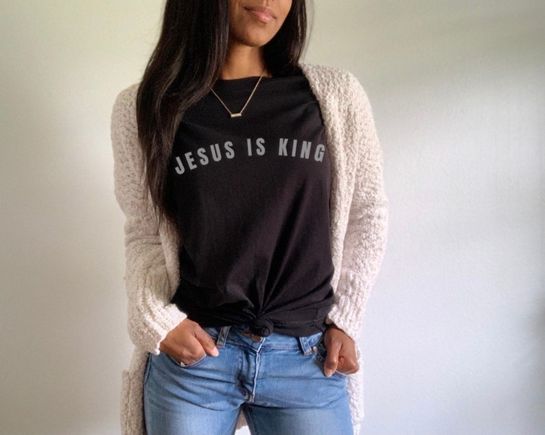 May include: A woman wearing a black t-shirt with the text "JESUS IS KING" in white lettering, a white cardigan, and blue jeans.