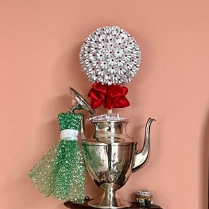 May include: A silver teapot with a white paper flower arrangement on top. The flowers are white with red centers and a red ribbon around the base. The teapot is on a wooden shelf with a small decorative box.