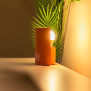 Op de afbeelding: Een moderne oranje tafellamp met een witte gloeilamp die aan staat. De lamp staat op een wit oppervlak.