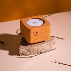 Puede incluir: Una vela cuadrada de color terracota con una etiqueta blanca que dice "SANDAL" y "Organic / Vegan 100% Soy Wax" y "TABART". La vela está sentada sobre una pequeña roca gris rugosa.