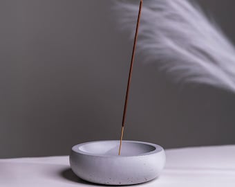 May include: A gray concrete incense holder with a brown incense stick burning and releasing white smoke.