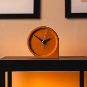 Può includere: Orologio analogico arancione con lancette nere e lancetta dei secondi argentata. L'orologio ha un design unico e arrotondato e si trova su una superficie di legno scuro. Lo sfondo presenta opere d'arte incorniciate.