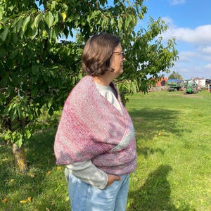 Könnte beinhalten: Ein rosa-grauer Strickschal ist über die Schultern einer Person gelegt. Der Schal hat ein strukturiertes Aussehen und wird über einem weißen Langarmshirt und einer blauen Jeans getragen. Der Hintergrund umfasst eine grüne Wiese, Bäume und einen blauen Himmel.