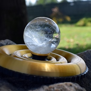Peut inclure: Une boule de cristal transparente est posée sur un socle doré en spirale avec un bord noir. La sphère présente des motifs internes. L'arrière-plan est une scène extérieure floue avec de l'herbe verte et des arbres.