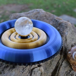 Può includere: Una scultura decorativa con una sfera di cristallo trasparente su anelli concentrici dorati e blu. È presente anche una conchiglia a spirale. La scultura è esposta all'aperto su una roccia.