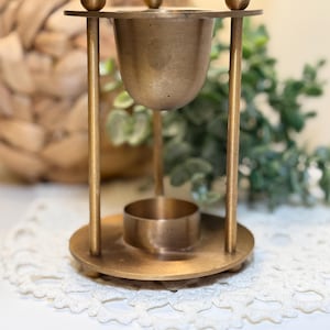 Peut inclure: Un brûle-parfum en métal doré composé d'une base circulaire avec un support à bougie central, trois tiges verticales surmontées de petites sphères et une coupelle suspendue au sommet pour les huiles ou les cires parfumées.