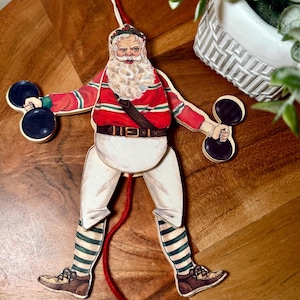 Könnte beinhalten: Ein Vintage-Weihnachtsschmuck mit dem Weihnachtsmann, der Hanteln hält. Der Holzausschnitt zeigt den Weihnachtsmann in einer rot-weiß gestreiften Jacke, einer weißen Hose und grün gestreiften Socken. Ein roter Faden mit einem roten Ball hängt von unten.