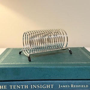 May include: A silver-toned metal wire rack featuring a series of connected circular loops arranged in a horizontal row. The rack is supported by four small legs, each tipped with a black protective cap, providing a stable base for the structure.