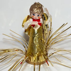 May include: A decorative angel figurine featuring a gold metallic cone-shaped dress, a white fuzzy collar with a red bow, and gold tinsel radiating from the base. The angel has light brown hair, a gold halo, and painted facial features.