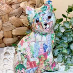 May include: A ceramic cat figurine featuring a colourful decoupage pattern of a garden scene with flowers, greenery, and architectural elements. The cat has pointed ears, dark eyes, and a small black nose, with a glossy finish covering the entire surface.