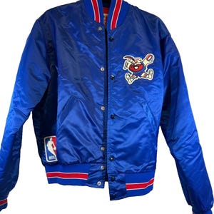 May include: A blue satin bomber jacket with red and white striped ribbing at the collar, cuffs, and hem. The jacket features a large embroidered logo of a cartoon character with a basketball on the left chest and a smaller NBA logo patch on the left side.