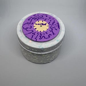 Op de afbeelding: Een kleine, ronde, zilveren glittertin met een paarse deksel. De deksel heeft een paars ontwerp met een geel midden. De tin wordt waarschijnlijk gebruikt voor opslag of als decoratief item.