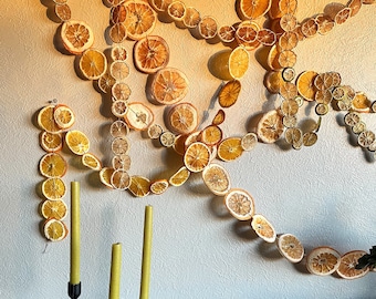 Może przedstawiać: Ściana ozdobiona girlandą z suszonych plastrów pomarańczy, z trzema czarnymi świecznikami i żółtymi świecami na drewnianym stole poniżej.