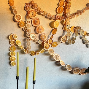 Multiple strings of dried citrus garlands, hanging artfully on a wall, with a trio of taper candles below on a wood surface