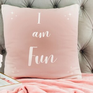 May include: A square, blush pink pillow with the words "I am Fun" in white script. The pillow has a soft, velvety texture and is decorated with small, white starburst designs. The pillow is resting on a pink blanket.