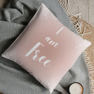 Puede incluir: Un cojín decorativo cuadrado con un diseño degradado rosa y blanco. El cojín presenta las palabras "I am Free" en escritura blanca. Pequeñas estrellas blancas están dispersas alrededor del texto. El cojín está sobre una manta gris y blanca.