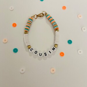 May include: A white beaded bracelet with turquoise and orange accents. The bracelet has the word "COUSIN" spelled out in black letters on white beads.