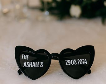 May include: Black heart-shaped sunglasses with white text. The left lens reads "THE ASHALL'S" and the right lens reads "29.08.2024". The sunglasses are on a white lace surface, with a blurred background of flowers and glassware.