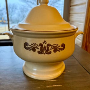 Puede incluir: Un tazón de servir de cerámica de color crema con un diseño floral marrón y una tapa. El tazón tiene una base de pedestal.