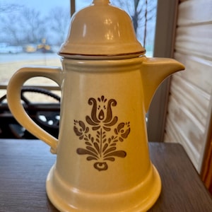 Puede incluir: Una cafetera de cerámica beige con tapa y asa. La cafetera presenta un diseño floral marrón en la parte delantera. El pico está en el lado derecho. Está sobre una superficie de madera.