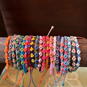 May include: A collection of colorful handmade bracelets displayed on a wooden dowel. The bracelets feature a floral design with various colors, including orange, green, blue, pink, and purple. Each bracelet has adjustable string ties.
