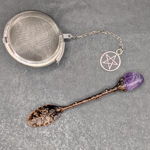 May include: A silver-toned metal tea infuser with a fine mesh sphere and a chain attached to a circular charm featuring a pentagram symbol, paired with a copper-toned metal spoon featuring a floral embossed bowl and a purple gemstone handle.