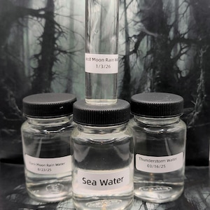 May include: A collection of four clear glass bottles filled with liquid. One tall thin bottle with a cork stopper labeled Wolf Moon Rain Water 1/3/26 sits atop a jar labeled Sea Water. Two other jars are labeled Black Moon Rain Water 8/23/25 and Thunderstorm Water 03/16/25.