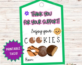 May include: Printable thank you tag with a green border and a pink and white text that says "Thank you for your support!" and "Enjoy your cookies". The tag features a cartoon cookie with a smiling face and three different types of Girl Scout cookies. The tag also includes the text "Printable Tags!" and "From:"