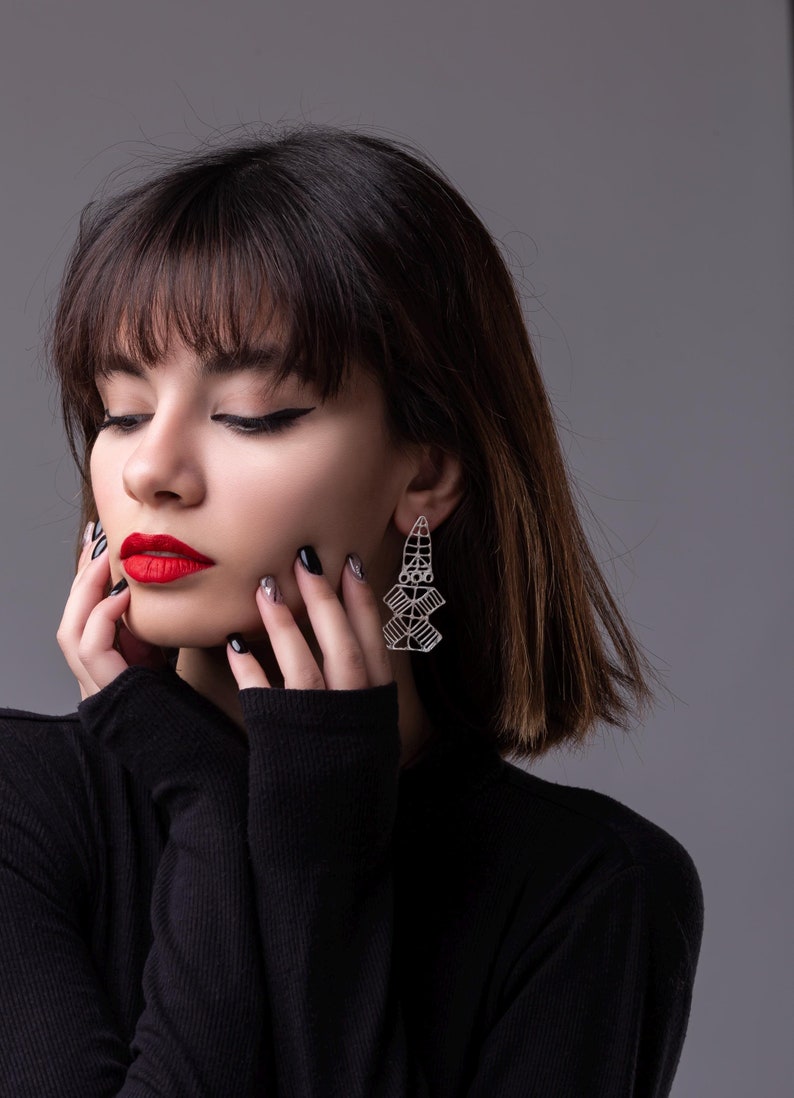 May include: A woman with dark brown hair and bangs wears a black turtleneck sweater. She is wearing silver geometric earrings. Her nails are painted black and silver.