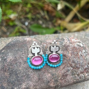 May include: A pair of silver earrings featuring a pink oval gemstone, encircled by small turquoise beads. The earrings have a decorative top. They are displayed on a textured rock surface.