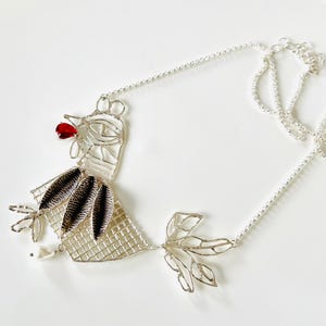 May include: A silver-toned wirework necklace in the shape of a bird. The bird has a red beak, black and white patterned feathers, and a leaf-shaped tail. The necklace hangs on a delicate chain.