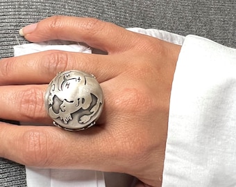 May include: A silver ring with a detailed design of a stylized face. The ring is worn on the index finger of a hand.