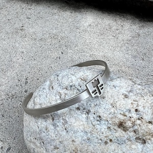 May include: A silver bracelet with a geometric design. The bracelet is on a gray rock.