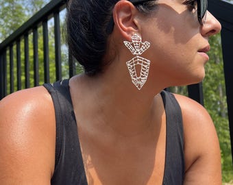 Könnte beinhalten: Eine Person trägt ein schwarzes ärmelloses Oberteil und große silberne Ohrringe mit geometrischem Design. Die Ohrringe sind in Form einer stilisierten Blume mit einer spitzen Unterseite geformt.