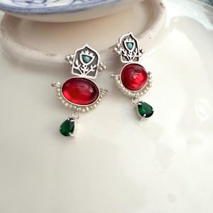 Puede incluir: Un par de pendientes de plata con una pieza metálica superior en forma de arco con una pequeña incrustación de turquesa, una piedra cabujón roja ovalada central, un borde de pequeñas perlas blancas y una gema verde en forma de lágrima.