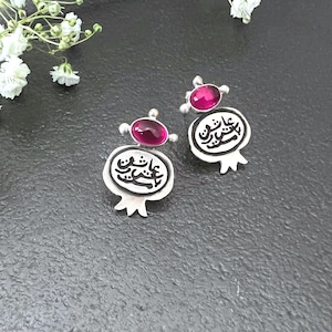 May include: A pair of silver-toned earrings with a pomegranate design. Each earring features a small, oval, magenta-colored stone at the top and a pomegranate shape below with Arabic script. The earrings are set against a dark background with white flowers.