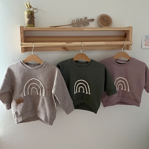 Könnte beinhalten: Drei Waffelstrick-Sweatshirts für Kinder mit Regenbogen-Stickerei. Die Sweatshirts sind in Beige, Olivgrün und Mauve.
