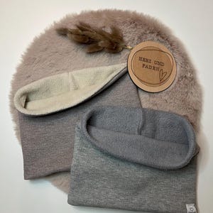 Könnte beinhalten: Zwei gerippte Strick-Halswärmer, einer in hellgrau und einer in dunkelgrau. Beide haben ein weiches Fleece-Futter. Die Halswärmer liegen auf einer hellbraunen Kunstfell-Oberfläche. Ein Holz-Etikett mit dem Text "Herz und Faden" ist im Hintergrund sichtbar.